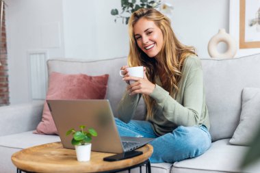 Evdeki bir kanepede oturup bir fincan kahve içerken dizüstü bilgisayarıyla çalışan güzel bir genç kadının fotoğrafı. Kameraya bakıyorum.