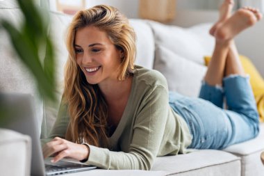 Evdeki kanepede yatarken dizüstü bilgisayarıyla çalışan güzel bir kadının fotoğrafı..