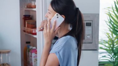 Evdeki mutfakta akıllı telefonla konuşurken buzdolabından sağlıklı yiyecekler alan güzel bir genç kadının videosu.