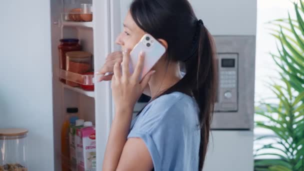 自宅のキッチンでスマートフォンと話しながら冷蔵庫から健康的な食べ物を取る美しい若い女性のビデオ — ストック動画