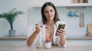 Evdeki mutfakta akıllı telefon kullanan güzel bir kadının yulaf lapası yerken videosu.