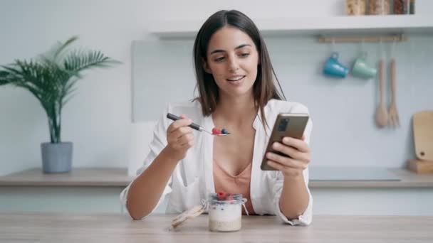 自宅のキッチンでスマートフォンを使用しながらベリーでお粥を食べる美しい女性のビデオ — ストック動画