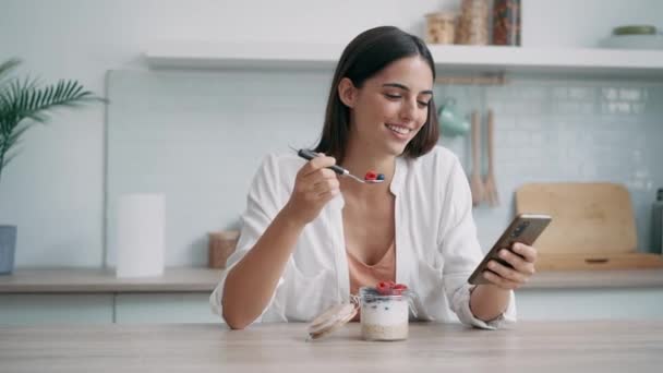 自宅のキッチンでスマートフォンを使用しながらベリーでお粥を食べる美しい女性のビデオ — ストック動画