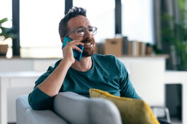 Gözlüklü, mutlu, olgun bir adamın evde kanepede dinlenirken akıllı telefonuyla konuşması..