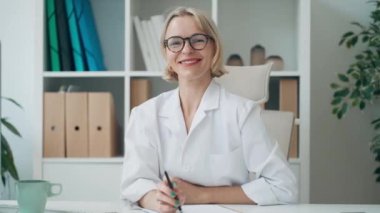 Konsültasyonda dizüstü bilgisayarla çalışırken kameraya gülümseyen olgun kadın doktorun videosu.