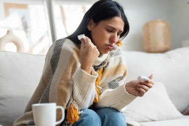 Evdeki kanepede battaniyeyle yatarken nezle olmuş, ateşi çıkan yorgun bir kadının fotoğrafı.