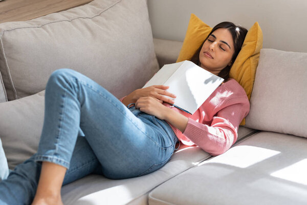 Shot of pretty young woman falling sleep while reading a book sitting on sofa at home
