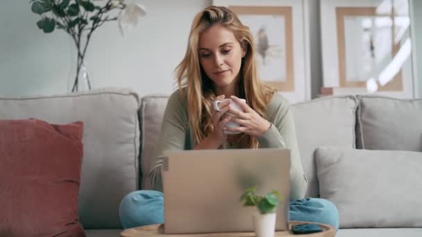 一个美丽的年轻女子坐在沙发上一边喝着咖啡一边用笔记本电脑工作的视频 — 图库视频影像