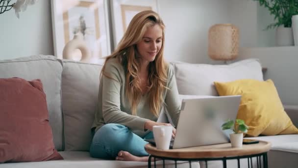 一个美丽的年轻女子坐在沙发上一边喝着咖啡一边用笔记本电脑工作的视频 — 图库视频影像