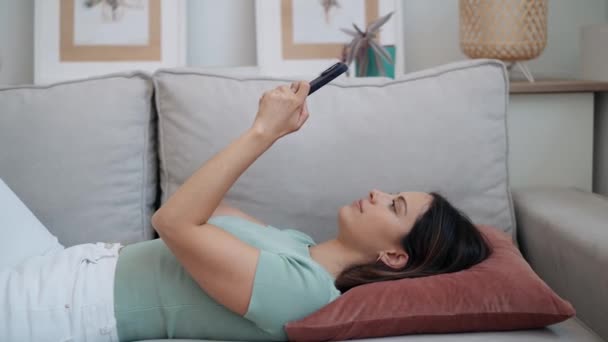 自宅でソファーに横たわっている間に携帯電話でメッセージを送信するかわいい若い女性のビデオ — ストック動画