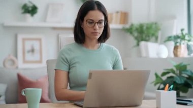 Video of concentrated beautiful business woman working with laptop while drinking coffee in living room at home.