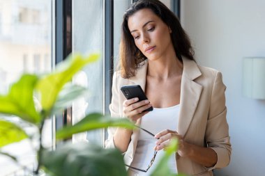 Ofisin penceresinin yanında cep telefonunu kullanan güzel bir kadının fotoğrafı.