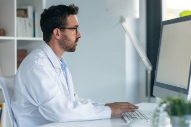 Tıbbi konsültasyonda notlar yazarken bilgisayarla çalışan olgun bir erkek doktorun fotoğrafı.