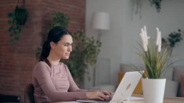 Video of concentrated beautiful business woman working with laptop while drinking coffee in living room at home.