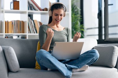 Evdeki bir kanepede dizüstü bilgisayarla çalışırken bir şeyi kutlayan mutlu bir kadının fotoğrafı..