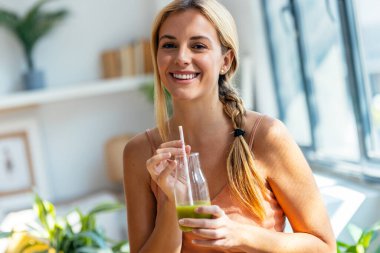 Evdeki oturma odasında kameraya bakarken sağlıklı yeşil smoothie içen mutlu kadının portresi..