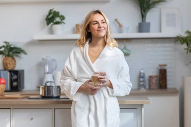 Bornozlu güzel bir kadının mutfakta ileriye bakarken bir fincan kahve içmesi.