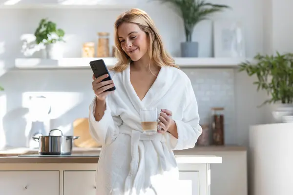 笑顔の若い女性が自宅のリビングルームで彼女の携帯電話を使用しながらコーヒーを楽しみます — ストック写真