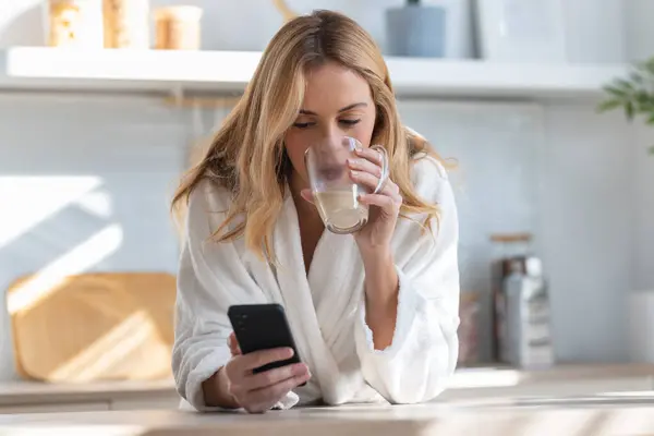 自宅のリビングルームで携帯電話を使用しながらコーヒーを楽しんでいる若い女性のショット — ストック写真