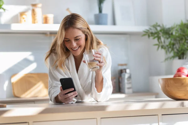 自宅のリビングルームで携帯電話を使ってコーヒーを楽しんでいる笑顔の女性のショット — ストック写真