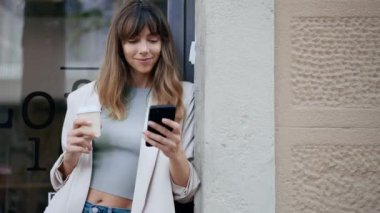 Güzel genç bir kadının elinde bir fincan kahveyle caddede dururken akıllı telefonunu kullanmasının videosu..