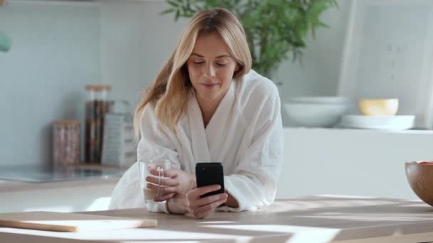 在家里客厅里 一个微笑的年轻女人一边用手机喝咖啡的视频 — 图库视频影像