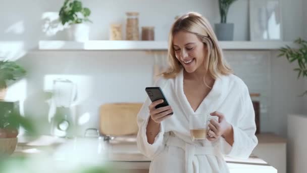 自宅のリビングルームで携帯電話を使ってコーヒーを楽しんでいる若い女性の笑顔のビデオ — ストック動画