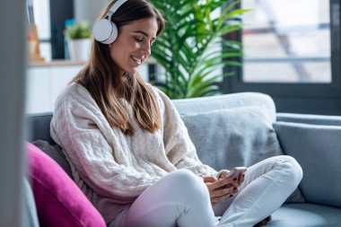 Evdeki kanepede otururken akıllı telefonuyla müzik dinleyen gülümseyen genç bir kadın..