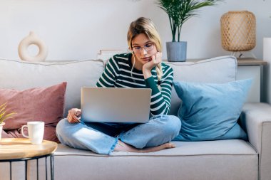 Evdeki kanepede otururken dizüstü bilgisayarıyla çalışan genç ve güzel bir kadının fotoğrafı..