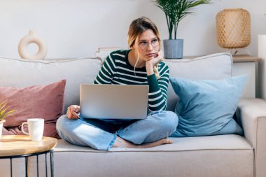 Evdeki kanepede otururken dizüstü bilgisayarıyla çalışan genç ve güzel bir kadının fotoğrafı..