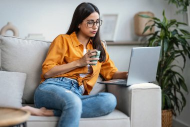 Evdeki bir kanepede oturup bir fincan kahve içerken dizüstü bilgisayarıyla çalışan kendine güvenen genç bir kadının fotoğrafı.