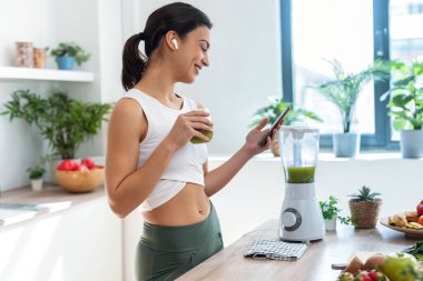 Shot of pretty woman using her mobile phone while drinking fruit detox juice in the kitchen at home.