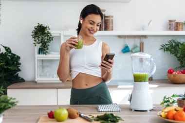 Shot of pretty woman using her mobile phone while drinking fruit detox juice in the kitchen at home.