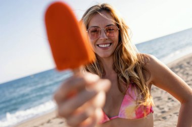 Yazın, bikinili, portakallı dondurma yiyen ve kameraya bakan çekici genç bir kadın.