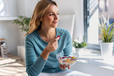 Evdeki mutfaktaki masada otururken sağlıklı meyve tabağı yiyen güzel bir kadının fotoğrafı.