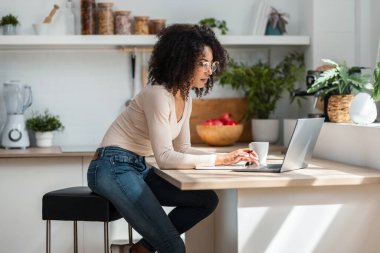 Evdeki mutfakta dizüstü bilgisayarla çalışan neşeli ve çekici bir kadın..