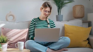 Evdeki kanepede otururken dizüstü bilgisayarıyla çalışan güzel bir genç kadının videosu..