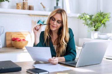 Evdeki mutfakta faturalara ve belgelere bakarken bilgisayarla çalışan genç ve güzel bir iş kadınının fotoğrafı..