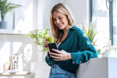 Evdeki mutfakta meyve suyu içerken güzel bir kadının cep telefonunu kullanması..