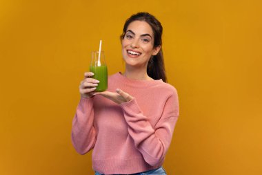 Çekici, gülümseyen, yeşil detoks suyu içen, izole edilmiş sarıda kameranın önünde poz veren bir kadın.