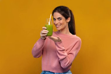 Çekici, gülümseyen, yeşil detoks suyu içen, izole edilmiş sarıda kameranın önünde poz veren bir kadın.