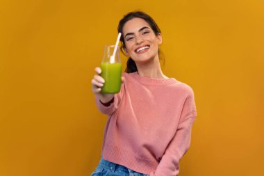 Çekici, gülümseyen, yeşil detoks suyu içen, izole edilmiş sarıda kameranın önünde poz veren bir kadın.