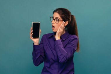 Shot of surprised attractive woman holding smartphone posing in front of camera on isolated blue clipart