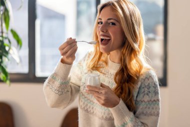 Evdeki oturma odasında dikilirken yoğurt yiyen mutlu bir kadın.