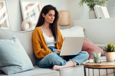 Evdeki kanepede otururken dizüstü bilgisayarıyla çalışan güzel bir genç kadının fotoğrafı..