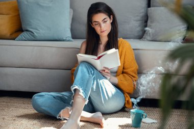 Güzel bir genç kadının kitap okuduğu ve evde yerde otururken bir fincan kahve içtiği bir fotoğraf.