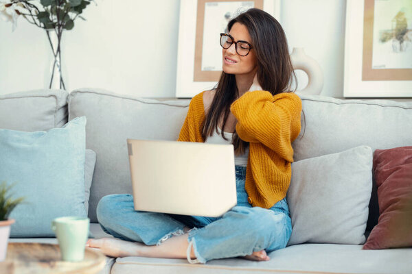 Уставшая молодая женщина с болью в шее, сидящая дома на диване с ноутбуком