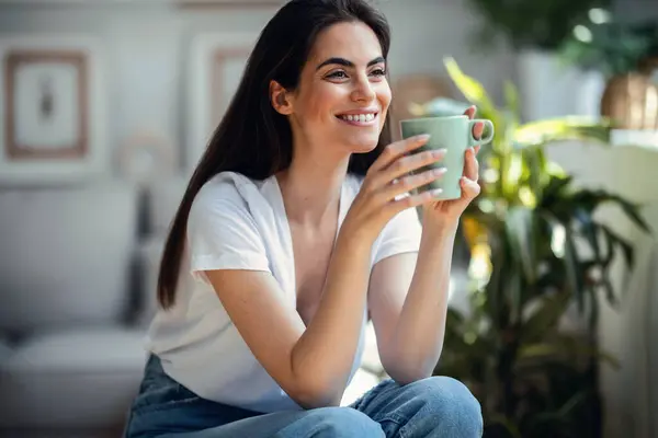 stock image Portrait of cute young woman posing at camera while drinking a cup of coffee in the living room at home.