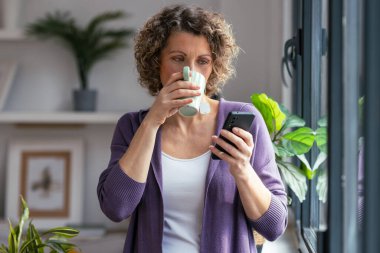 Evdeki oturma odasının penceresinin yanında kahve içerken güzel olgun bir kadının cep telefonunu kullanması.