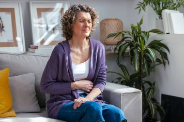 Shot of unhealthy mature woman with stomachache leaning on the couch in the living room at home clipart